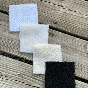 May include: Four fabric swatches, three white and one black, with a fuzzy texture. The swatches are arranged in a vertical stack on a wooden surface.