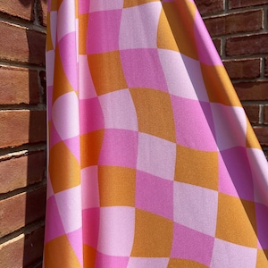 May include: A pink and orange checkered fabric with a wavy pattern. The fabric is folded and draped over a brick wall.