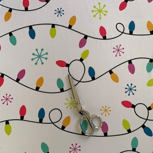 May include: White fabric with a repeating pattern of colorful lightbulbs and snowflakes. A pair of silver scissors is lying on the fabric.