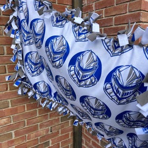 May include: White fleece blanket with a repeating pattern of blue and gray knight helmets. The blanket has a knotted fringe in gray and white. The blanket is draped over a metal pole and in front of a brick wall.