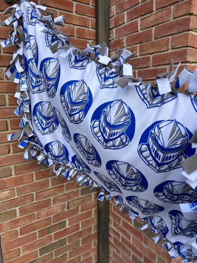 May include: White fleece blanket with a repeating pattern of blue and gray knight helmets. The blanket has a knotted fringe in gray and white. The blanket is draped over a metal pole and in front of a brick wall.