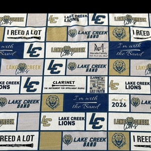 May include: Fabric with a repeating pattern of navy blue, gold, and white squares. Each square features text and graphics related to the Lake Creek Lions band, including the school logo, clarinets, and the phrase "I'm with the Band."