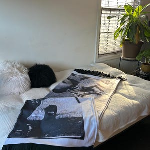 May include: Two black and white decorative throw blankets with fringe are displayed on a bed. The blankets feature a black and white image and are folded. A white bedspread and two decorative pillows are also visible.