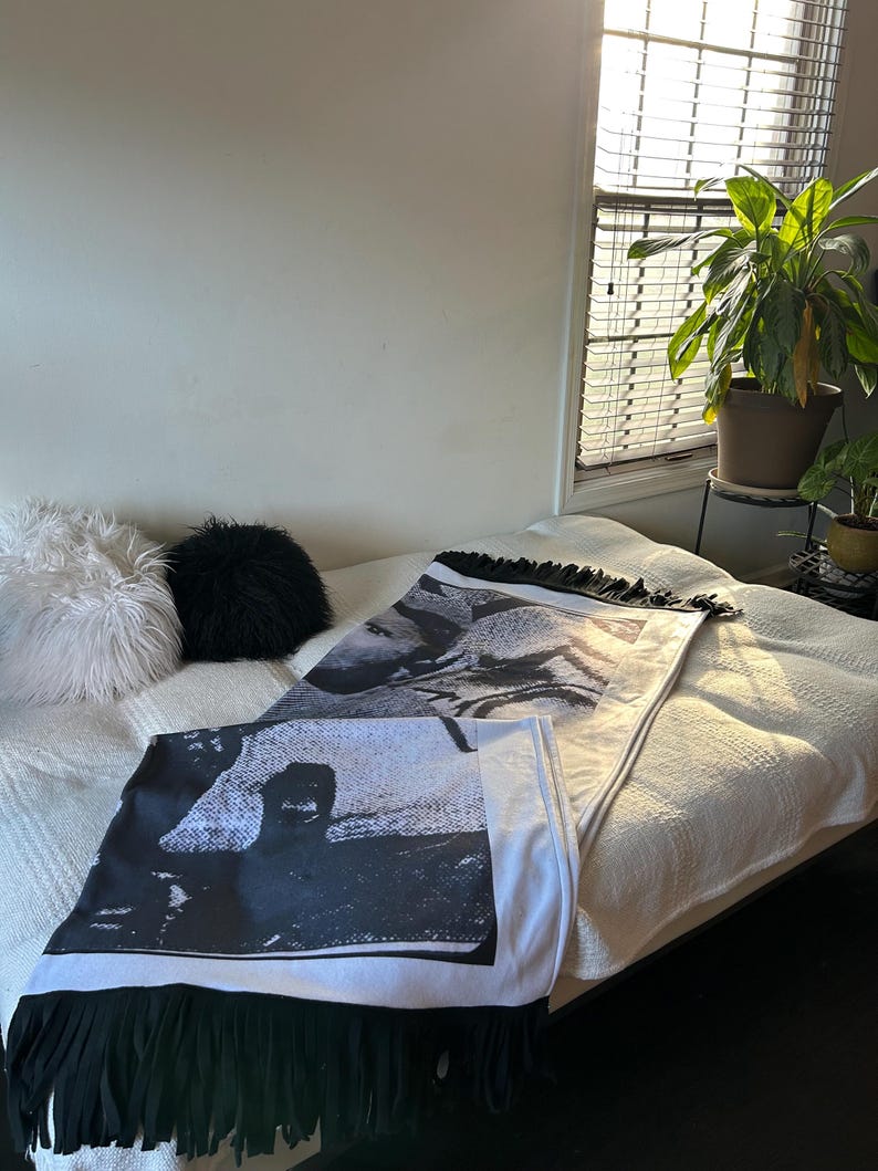 May include: Two black and white decorative throw blankets with fringe are displayed on a bed. The blankets feature a black and white image and are folded. A white bedspread and two decorative pillows are also visible.