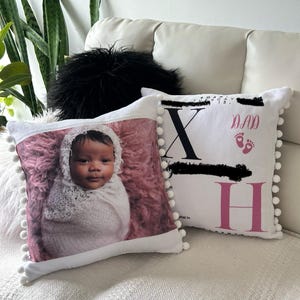 May include: Two white decorative pillows with pom-pom trim. One pillow features a baby wrapped in a white blanket on a pink background. The other pillow has the letters "X" and "H" with the word "DAD" and footprints.