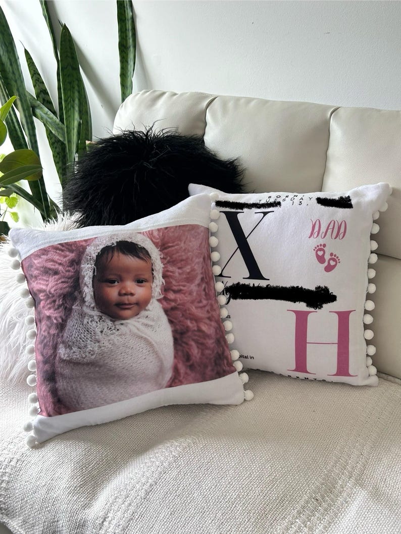 May include: Two white decorative pillows with pom-pom trim. One pillow features a baby wrapped in a white blanket on a pink background. The other pillow has the letters "X" and "H" with the word "DAD" and footprints.