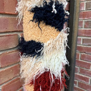 May include: A textured, rectangular wall hanging with a shaggy, multi-colored design. The piece features sections of cream, tan, navy, red, and pink, creating a unique, abstract pattern. The fibers are long and create a soft, tactile appearance.