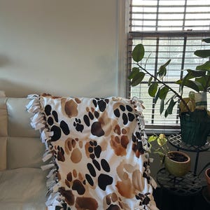 May include: A white blanket with a repeating pattern of brown and black paw prints. The blanket has a white fringe border. The blanket is draped over a white couch.