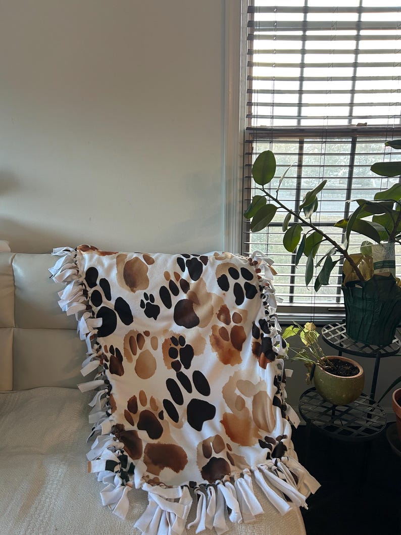 May include: A white blanket with a repeating pattern of brown and black paw prints. The blanket has a white fringe border. The blanket is draped over a white couch.