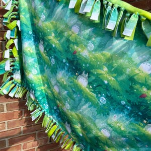 May include: A green and blue fleece blanket featuring a forest scene with cartoon characters. The blanket has a fringe border of green and white fabric strips. The blanket is displayed against a brick wall.