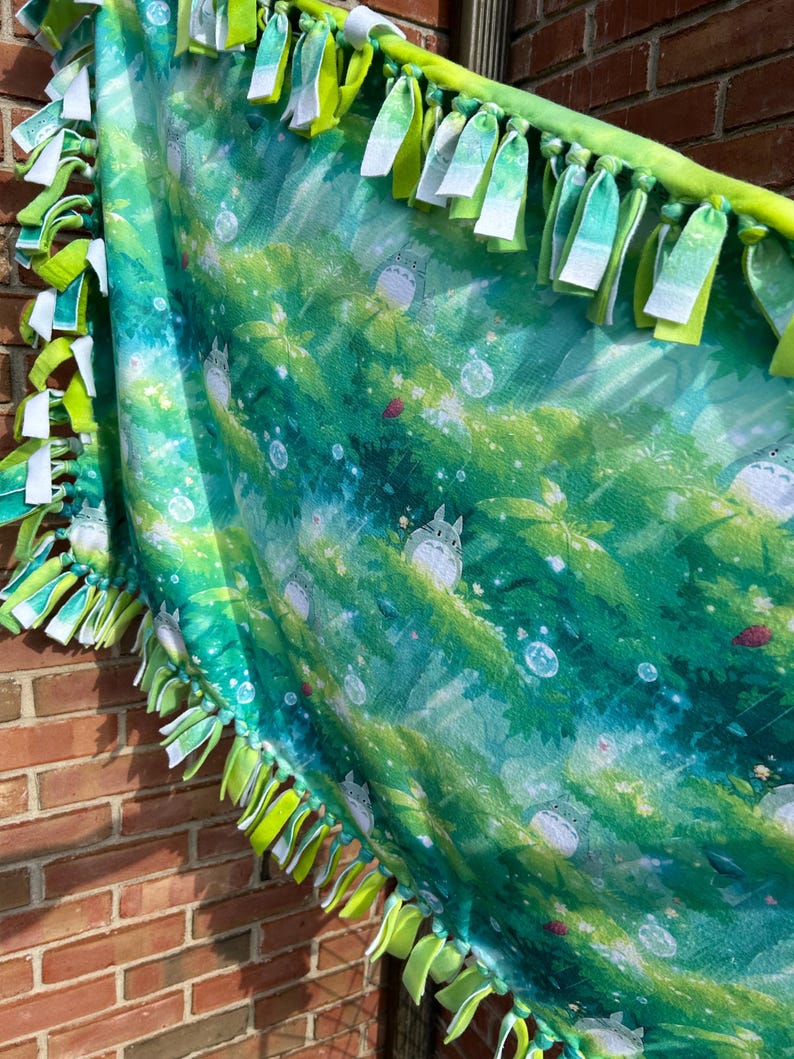 May include: A green and blue fleece blanket featuring a forest scene with cartoon characters. The blanket has a fringe border of green and white fabric strips. The blanket is displayed against a brick wall.