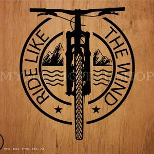May include: Black graphic of a mountain bike inside a circle with the text "RIDE LIKE THE WIND". The design includes mountains, water, and stars. The image is on a wood grain background.