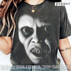 Puede incluir: Una camiseta de manga corta gris carbón con un gran estampado gráfico en blanco y negro de un rostro humano con una expresión intensa y distorsionada. La palabra pepper está impresa en la esquina superior derecha de la prenda.