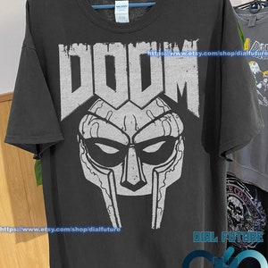 Op de afbeelding: Zwart T-shirt met een witte afbeelding van het masker van rapper MF DOOM. De tekst "DOOM" staat boven het masker. De tekst "https://www.etsy.com/shop/dialfuture" staat onder het masker. De tekst "DIAL FUTURE" staat in de rechterbenedenhoek van de afbeelding.