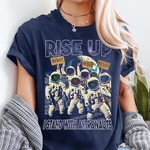Puede incluir: Una camiseta azul marino con el texto "Rise Up" en letras blancas. La camiseta presenta un gráfico de seis astronautas con trajes espaciales que sostienen carteles que dicen "Resist". El texto "I Stand With Astronauts" está impreso debajo del gráfico.