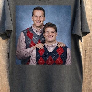 May include: Dark gray t-shirt featuring a printed image of two men wearing argyle vests over button-down shirts. The image has a blue background. The t-shirt is hanging on a wooden hanger.