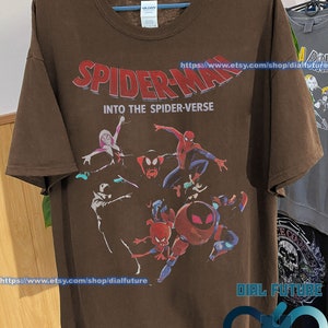 Pode incluir: Camiseta marrom com uma estampa gr&aacute;fica dos personagens do Homem-Aranha do filme "Spider-Man: Into the Spider-Verse". O texto "Spider-Man: Into the Spider-Verse" est&aacute; impresso em vermelho na camiseta. A camiseta tem gola redonda e mangas curtas.