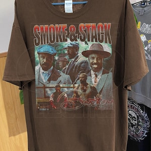 Puede incluir: Camiseta marrón con un diseño gráfico de estilo vintage. El texto "SMOKE & STACK" está sobre un collage de hombres con trajes y sombreros. La palabra "Brothers" también es visible. La camiseta está colgada en una percha.