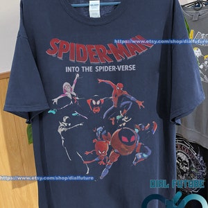 Pode incluir: Uma camiseta azul marinho com uma estampa gr&aacute;fica dos personagens do Homem-Aranha do filme "Spider-Man: Into the Spider-Verse". O texto "Spider-Man: Into the Spider-Verse" est&aacute; impresso em letras vermelhas e brancas na camiseta.