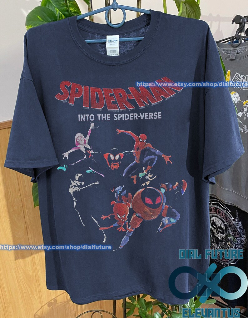Pode incluir: Uma camiseta azul marinho com uma estampa gr&aacute;fica dos personagens do Homem-Aranha do filme "Spider-Man: Into the Spider-Verse". O texto "Spider-Man: Into the Spider-Verse" est&aacute; impresso em letras vermelhas e brancas na camiseta.