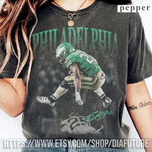 Peut inclure: Un t-shirt gris anthracite à manches courtes avec un graphisme représentant un joueur de football américain en uniforme vert et le texte PHILADELPHIA arqué au-dessus. Le motif inclut une image plus petite avec le numéro 26 et une signature verte.