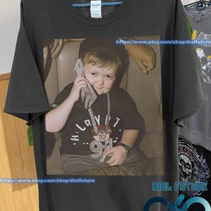 Peut inclure: Un t-shirt noir avec une photo d'un jeune garçon portant une chaîne en or et un sweat-shirt noir. Le garçon tient un paquet d'argent à son oreille et regarde la caméra. Le texte "https://www.etsy.com/shop/dialfuture" est imprimé en bas du t-shirt.