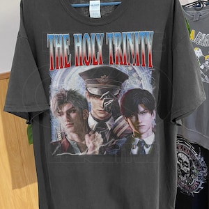 Könnte beinhalten: Dunkelgraues T-Shirt mit einer Grafik von drei Männern und dem Text "THE HOLY TRINITY" in Rot und Weiß. Das Design zeigt einen Mann mit Hut im Militärstil und zwei weitere Männer.