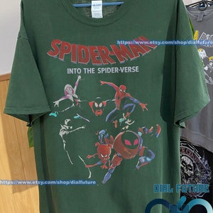 Pode incluir: Camiseta verde com uma estampa gr&aacute;fica dos personagens do Homem-Aranha do filme "Spider-Man: Into the Spider-Verse". O texto "Spider-Man: Into the Spider-Verse" est&aacute; impresso em letras vermelhas e brancas na camiseta. A camiseta tem gola redonda e mangas curtas.