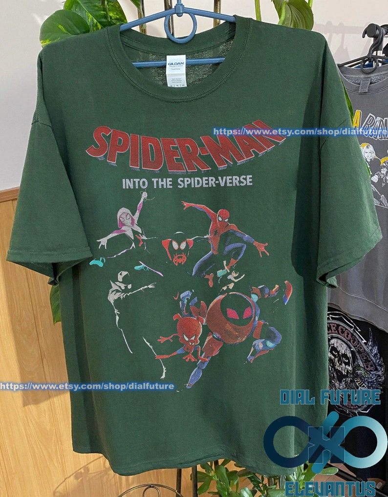 Pode incluir: Camiseta verde com uma estampa gr&aacute;fica dos personagens do Homem-Aranha do filme "Spider-Man: Into the Spider-Verse". O texto "Spider-Man: Into the Spider-Verse" est&aacute; impresso em letras vermelhas e brancas na camiseta. A camiseta tem gola redonda e mangas curtas.