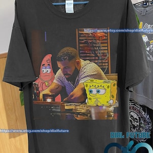 May include: Black t-shirt with a graphic print of Drake, Spongebob Squarepants, and Patrick Star. The text "https://www.etsy.com/shop/dialfuture" is printed on the bottom of the shirt.