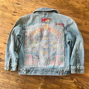 May include: Light blue denim jacket with a Guns N' Roses graphic on the back. The graphic features a cross, skulls, and the band's name. The jacket has button closures and a collared neckline. The graphic is stitched onto the jacket.