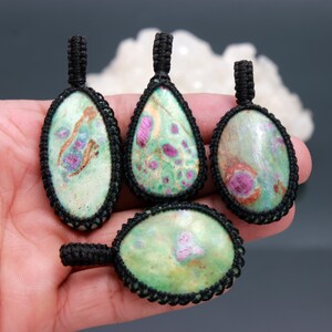 May include: Four pendants featuring polished green and pink stones encased in black macrame cord. Each stone displays a unique pattern of light green, dark green, and vibrant pink inclusions. The pendants are shaped as ovals and a teardrop.