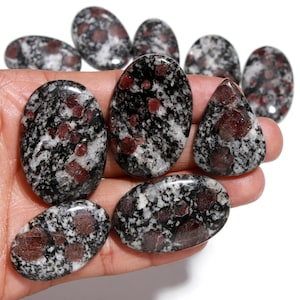 May include: A collection of polished gemstones with a speckled black and white pattern, interspersed with deep red inclusions. The stones vary in shape, including oval and teardrop forms. The gemstones are held in a hand.
