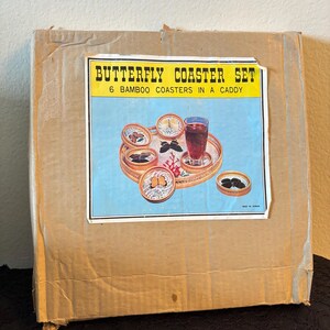 Peut inclure: Une bo&icirc;te en carton avec une &eacute;tiquette indiquant "BUTTERFLY COASTER SET". L'&eacute;tiquette montre six dessous de verre en bambou dans un support, avec un fond bleu et une boisson sur un plateau. La bo&icirc;te est marron et usag&eacute;e.
