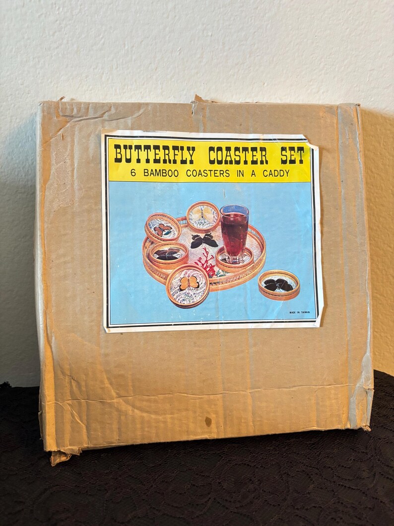 Peut inclure: Une bo&icirc;te en carton avec une &eacute;tiquette indiquant "BUTTERFLY COASTER SET". L'&eacute;tiquette montre six dessous de verre en bambou dans un support, avec un fond bleu et une boisson sur un plateau. La bo&icirc;te est marron et usag&eacute;e.