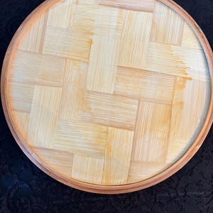 Peut inclure: Plateau rond en bambou tress&eacute;, avec un bord marron clair et une surface tiss&eacute;e naturelle de couleur claire. Le design du plateau pr&eacute;sente un motif de tissage en panier, mettant en valeur le grain naturel du bois.