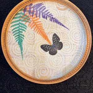 Peut inclure: Plateau d&eacute;coratif rond avec un cadre en bois naturel. Le plateau pr&eacute;sente un dessus en verre avec un motif de feuilles de foug&egrave;res color&eacute;es en vert, orange et violet, ainsi qu'un papillon noir et blanc. Le fond a un motif cr&egrave;me.