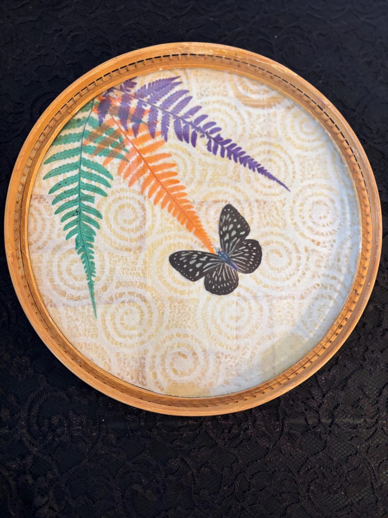 Peut inclure: Plateau d&eacute;coratif rond avec un cadre en bois naturel. Le plateau pr&eacute;sente un dessus en verre avec un motif de feuilles de foug&egrave;res color&eacute;es en vert, orange et violet, ainsi qu'un papillon noir et blanc. Le fond a un motif cr&egrave;me.