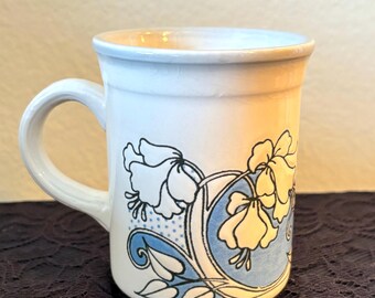 Peut inclure: Une tasse en céramique blanche ornée d'une illustration décorative de fleurs jaunes et bleues avec des feuilles vertes. Le motif comprend un élément de fond bleu circulaire et des contours noirs, avec une anse incurvée sur le côté gauche.