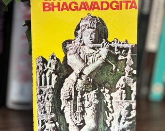 Peut inclure: Un livre de poche intitulé The Bhagavadgita, transcréé par P. Lal. La couverture présente un fond jaune vif avec un texte rouge en gras et une illustration en relief en pierre monochrome détaillée d'une figure jouant de la flûte, entourée de sculptures.