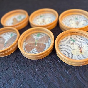 Peut inclure: Six dessous de verre ronds en bois avec un dessus en verre, chacun pr&eacute;sentant un papillon pr&eacute;serv&eacute;. Les papillons sont de couleurs vari&eacute;es, avec des nuances de marron, de blanc et de jaune. Les dessous de verre sont dispos&eacute;s sur une surface sombre et textur&eacute;e.