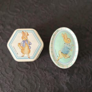 May include: Two vintage Peter Rabbit themed tins. One is hexagonal with a blue and white border, and the other is oval. Both feature illustrations of Peter Rabbit in a blue jacket.