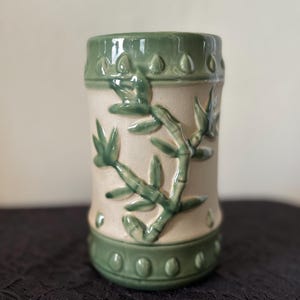 May include: A ceramic mug with a bamboo design. The mug is cylindrical with a light beige center and green bands at the top and bottom. Green bamboo stalks and leaves are sculpted on the mug's surface. The mug has a glossy finish.
