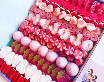 Puede incluir: Una caja de regalo de dulces rosa y roja llena de varios dulces, incluyendo corazones de gominola, ositos de gominola y malvaviscos. La caja está decorada con el texto "The Sweet Life".