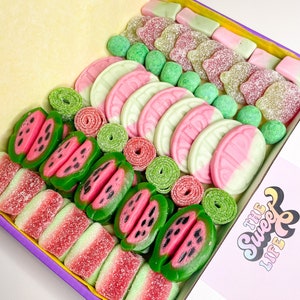 May include: A purple box filled with assorted gummy sweets. The sweets are shaped like watermelon slices, green and pink swirls, and green balls. The box is open and the sweets are visible. The box has a pink label with the text "The Sweet Life" in white.