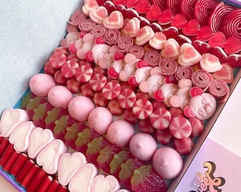 Puede incluir: Una caja de regalo de dulces rosa y roja llena de varios tipos de gomitas, malvaviscos y otros dulces. La caja está decorada con el texto "The Sweet Life".
