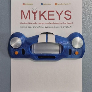 May include: A blue 3D-printed key rack in the shape of a vintage car. It has silver headlights, orange indicators, and white racing stripes. The text "MYKEYS" is displayed above the car, with product information below.