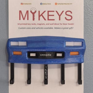 May include: Blue 3D-printed key rack shaped like a car front, with the text "CAMARO". It has four black hooks for keys and a white background with the text "MY KEYS".