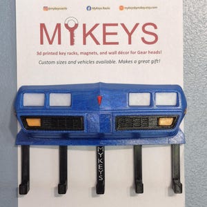 May include: A blue 3D-printed key rack shaped like a car front, with four key hooks. The rack has white rectangular details and orange-yellow lights. The text "MYKEYS" is visible on the rack and the backing card.