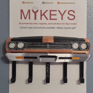 May include: 3D-printed key rack shaped like a classic car front end. The rack is black, copper, and silver, with four black hooks for keys. The sign above reads "MYKEYS" and mentions custom sizes.